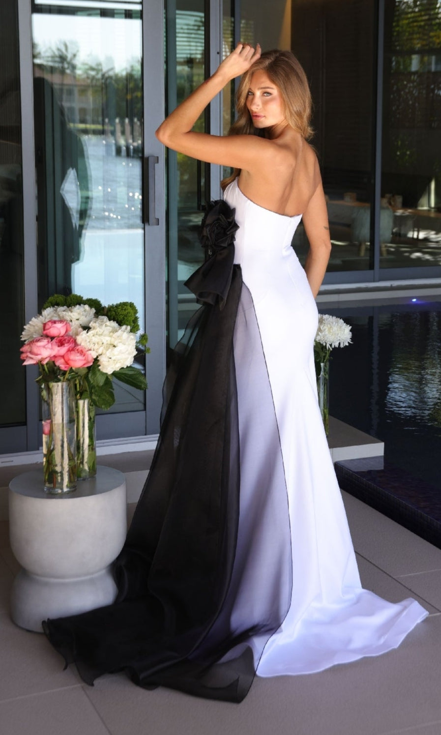 A woman in the Ava Presley 26518 long prom dress, featuring a strapless design with a large black organza bow and train, stands by a glass door near floral arrangements and a pool.